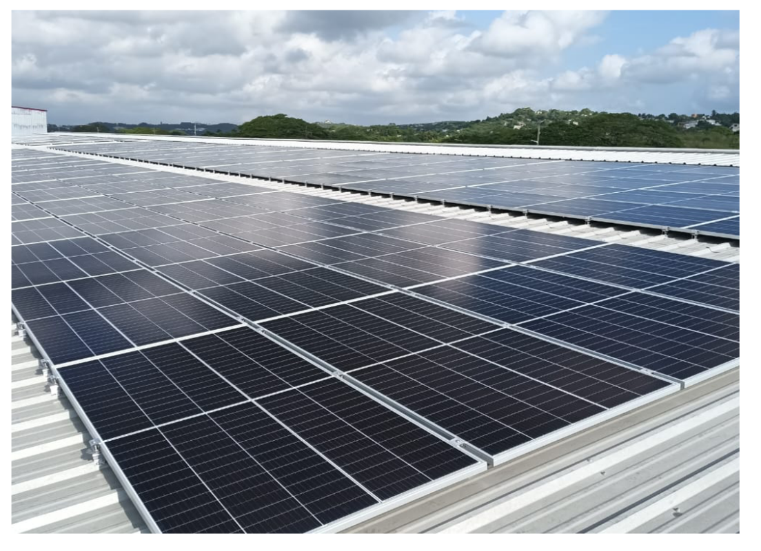 A rooftop covered with solar panels under a partly cloudy sky with trees and hills in the distant background.